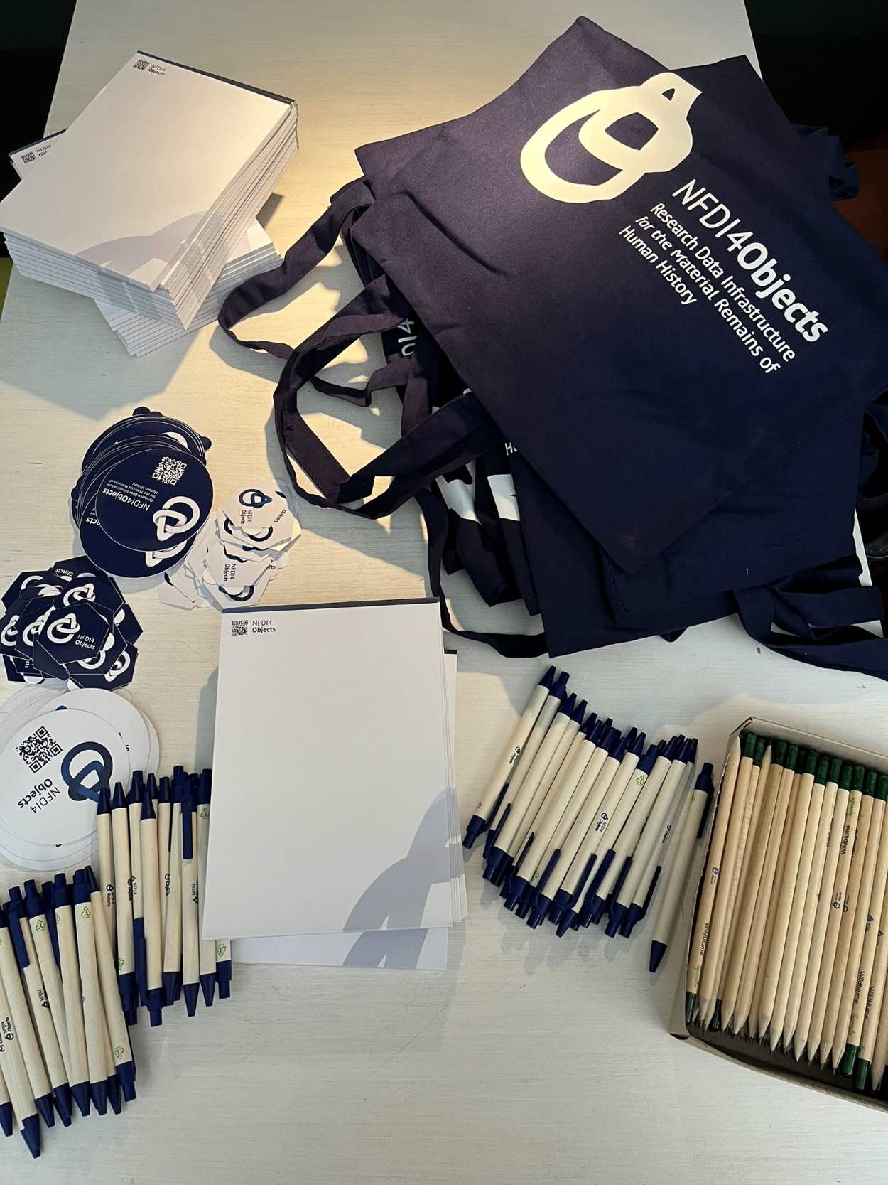 Für alle Teilnehmenden gab es ausreichend NFDI4Objects Sticker, Schreibblöcke, Kugelschreiber, Bleistifte und Stoffbeutel zum Mitnehmen. Merchandise-Produkte von NFDI4Objects liegen auf einem Tisch aus.