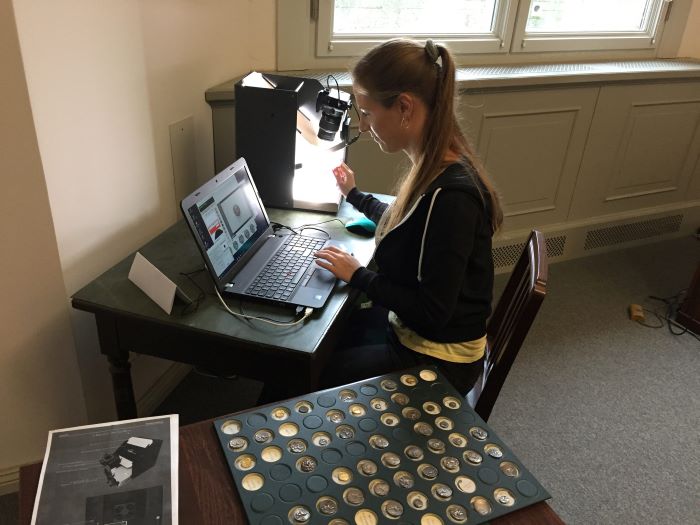 The photo shows a woman working at a desk. She is examining a coin with an apparatus and entering data into a laptop.